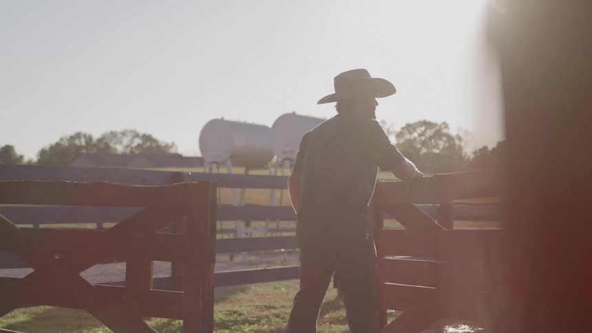 A person wearing a widebrimmed hat and overalls stands on sunlit dirt opens a wooden gate walks through the opening and is strongly backlit against a hazefilled landscape with distant rounded structures