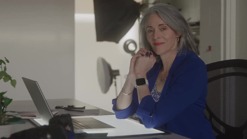 A confident gray haired woman wearing a blue blazer sits at a brightly lit desk with a laptop and camera equipment turning to smile at the camera