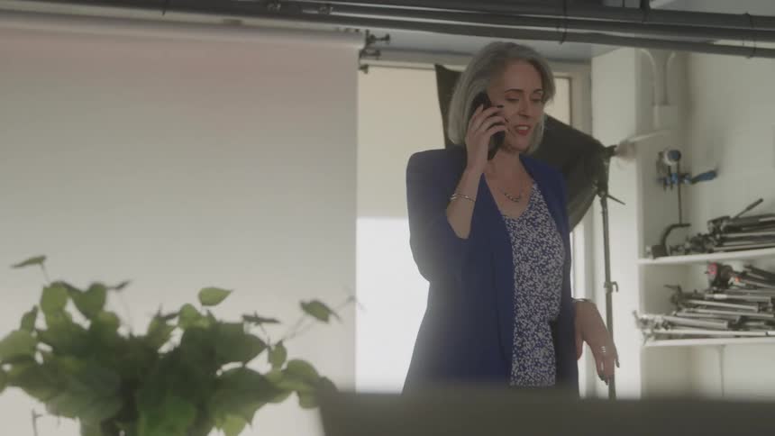 A grayhaired woman wearing a blue jacket stands in an interior room talks on a mobile telephone held to her ear gestures with her free hand and stands near a large light fixture and shelves of equipment with a leafy green plant in the foreground