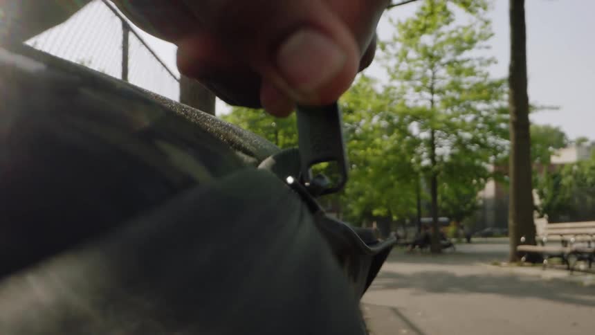 A persons hand unzips a black material container in the foreground revealing a sunny park setting with trees black benches and a wire fence in the background