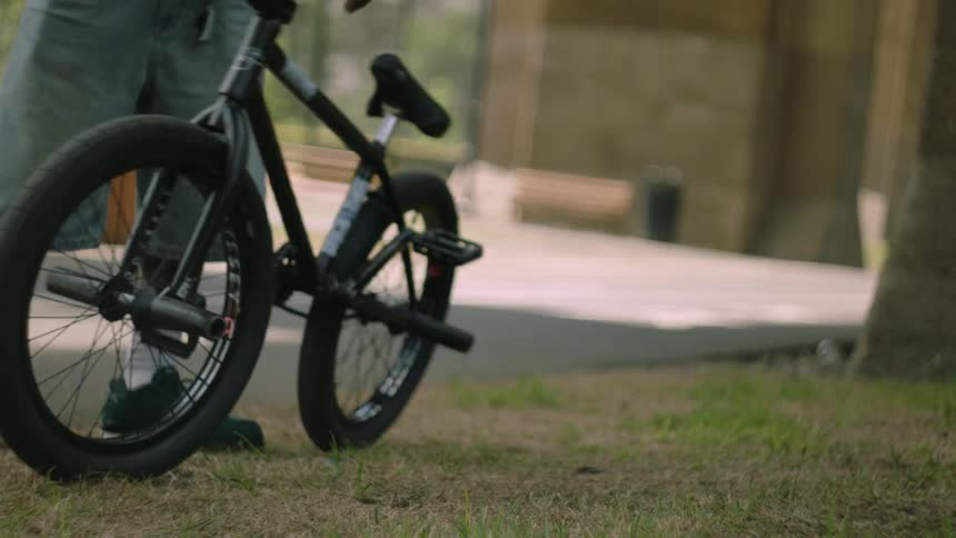 The video shows a bmx bicycle lying on its side in a park on a patch of grassa person in a lightcolored denim shorts and black sneakers walks past the bike and continues out of the frame