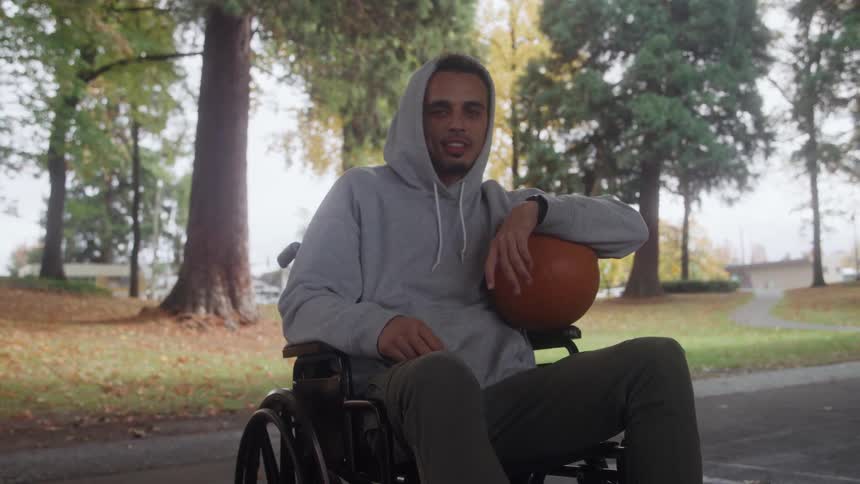 A young man in a wheelchair wearing a gray coat and holding a basketball looking forward