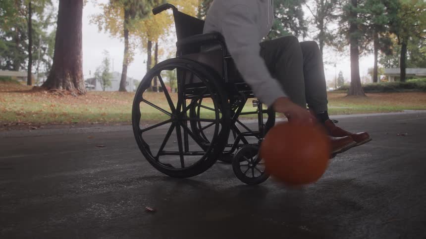 A close view of the lower body shows a man in a wheelchair wearing a grey hoodie and brown boots dribbling an orange basketball on a paved area in an autumn park