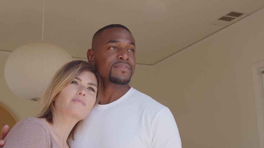 A man and a woman stand close together indoors in a brightly lit room the woman rests her head against the mans shoulder the man has an arm around the woman and both look upward and away from the camera