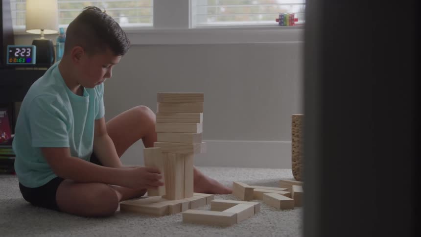 A young boy in a teal shirt sits on a carpet building a tall wooden block tower beside a woven basket in a bright room
