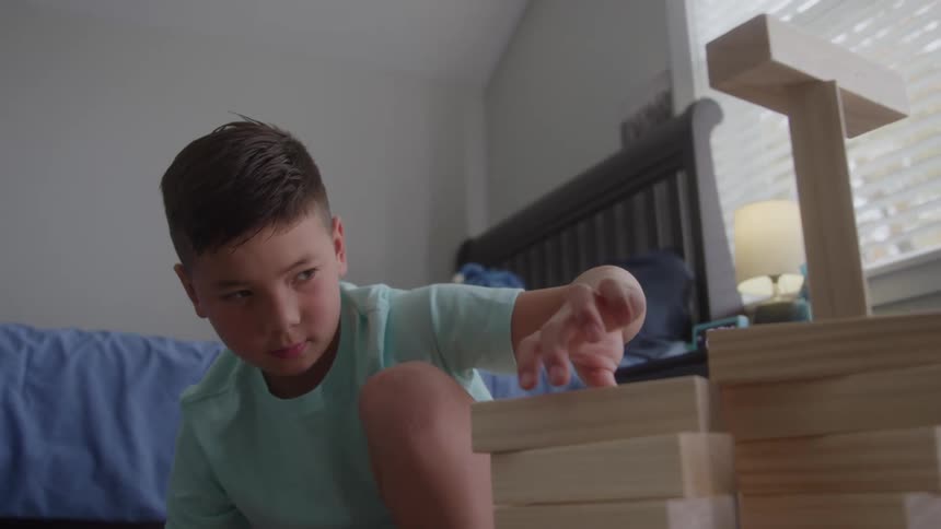 A young boy wearing a light blue shirt kneels on the floor in a bright bedroom concentrating intently as he carefully places a large wooden block on a tall stack