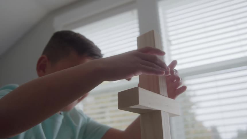 The boy is building a structure with wooden blocks near a window appearing happy and concentrating on his work