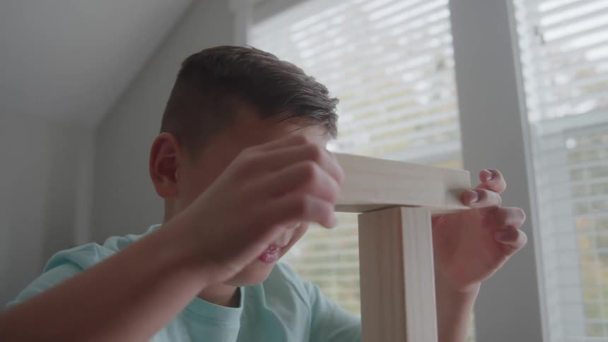 The video is an interior shot of a young boy in a light blue shirt looking up intently as he plays with wooden building blockshe is seated near a large window with blinds and the scene suggests he is in a brightly lit bedroom or play area
