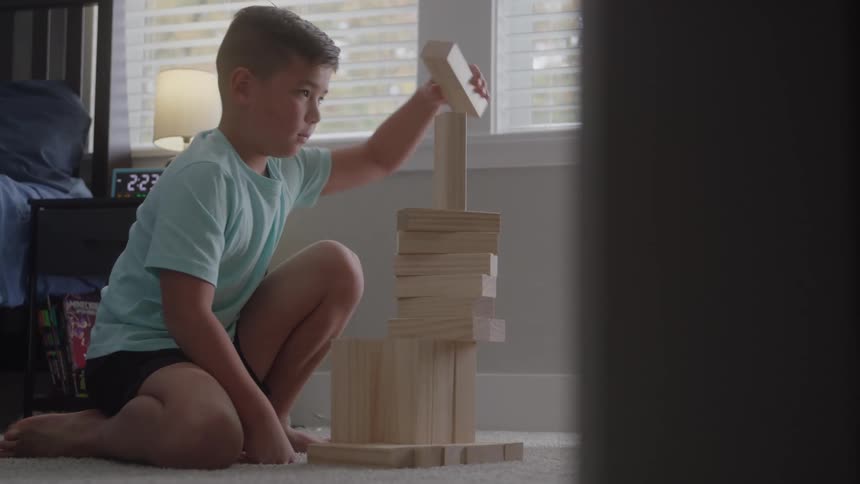 A young boy in a light blue shirt and black shorts kneels on the carpet and intently plays a stacking game with large wooden blocks in a bright bedroom