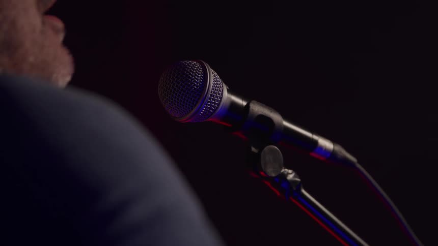 A close side view from behind shows an older man with a beard leaning in to sing into a dark microphone on a stand against a dim stage background with subtle red light