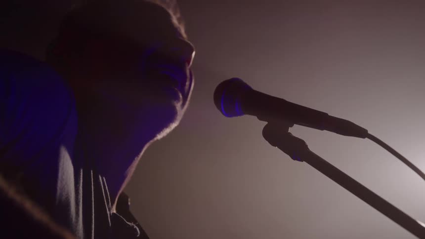 A person sings into a microphone with his profile silhouetted against a bright hazy light the microphone angled toward his mouth in an indoor setting
