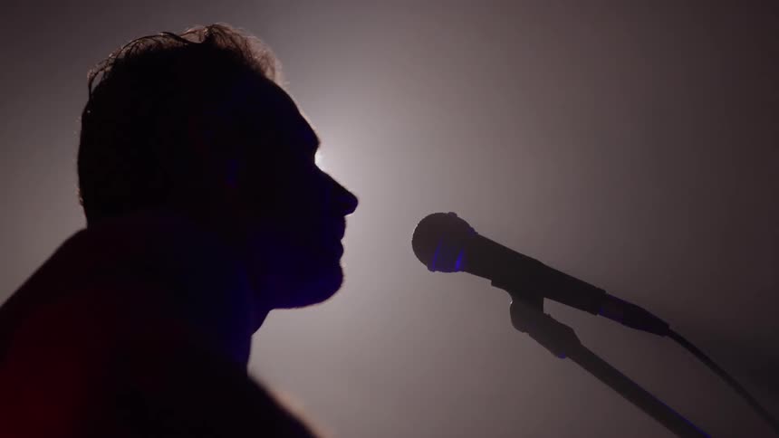 The silhouette of a man singing into the microphone and a dark room with a white light in the background