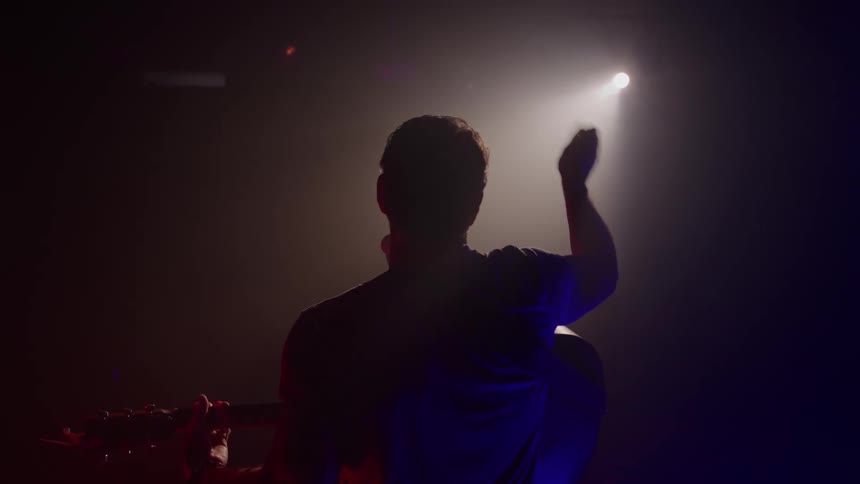 A silhouette of a male musician playing a guitar faces a bright spotlight on a dark stage lit with red and blue stage lights raising his hand to gesture