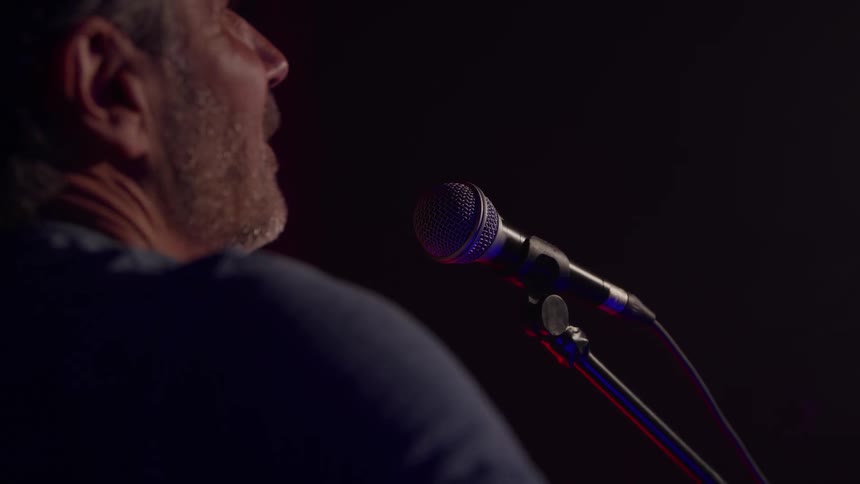 A close side view shows an older man with a beard singing into a dark microphone on a stand in a dimly lit performance space