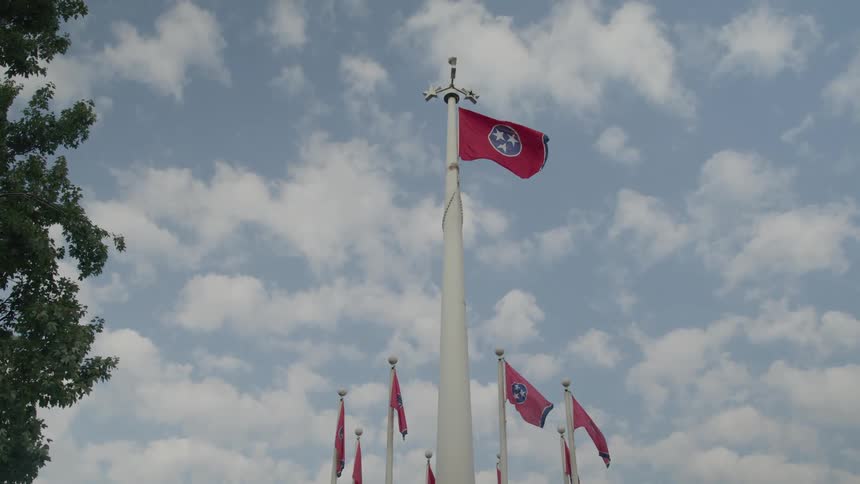 The bright red flag of tennessee on a tall pole snaps in the wind surrounded by several smaller flags on a sunny day with a blue sky and white cumulus clouds