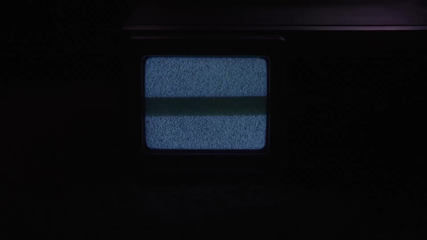 A medium shot of a small square monitor in a dark room displays a blank blue screen that suddenly breaks into flickering white and gray static