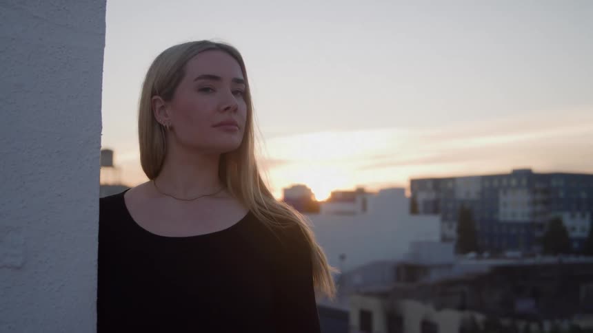 A woman with long blonde hair wearing a black top stands by a white wall gazing toward the sunset over a city skyline