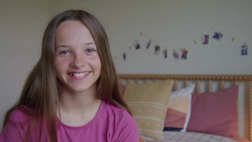 The video is a mediumcloseup shot of a young girl with long brown hair and bright blue eyes smiling at the camerashe is sitting in a sunlit bedroom in front of a bed with a wooden headboard and a string of photos is visible on the wall behi