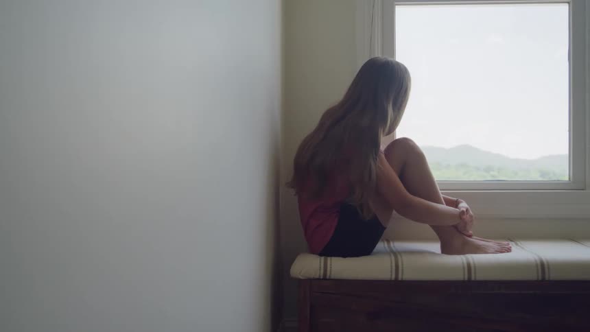 A barefoot long haired girl wearing a dark shirt and shorts sits with her knees pulled up on a window seat looking out at a bright green mountainous landscape