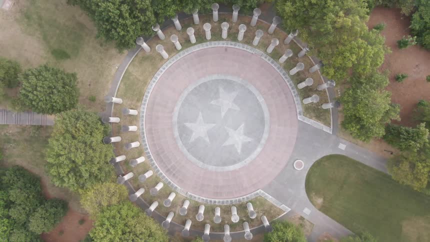 A top down aerial view slowly zooms out to reveal a large circular monument with the tennessee tristar in the center surrounded by a ring of columns and set within a lush green park