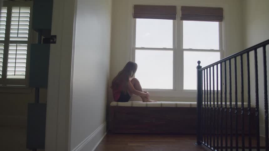 The video is an interior shot of a hallway where a woman sits on a window bench looking outsidethe bright sunlight streaming through the windows silhouettes her form against the dark hallway and black wrought iron railing