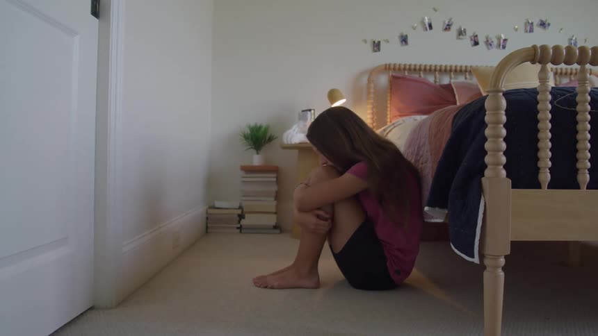 A barefoot long haired girl wearing a pink shirt and black shorts sits huddled on the floor with her knees to her chest next to a bed in a bright bedroom