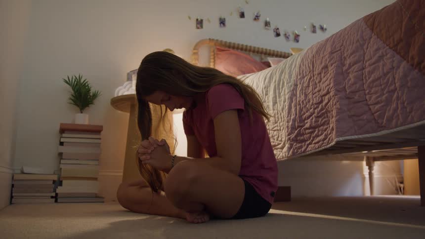 A teenage girl sits on a carpeted floor with her legs crossed her hands clasped together her head bowed in a bedroom in a prayer position and a bed and nightstand filling the background