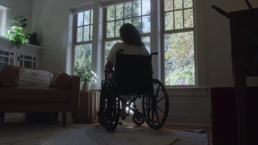 Woman sitting in a wheelchair in the living room stares out the window on an autumn day