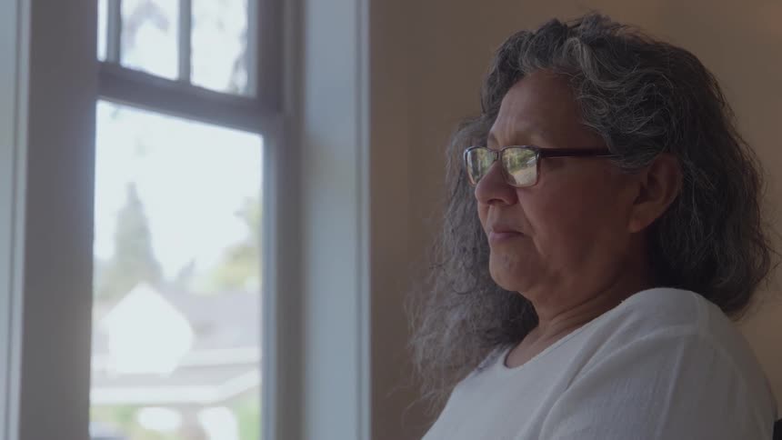 A close side view shows an older woman with gray curly hair and eyeglasses wearing a white shirt and looking out a window on a bright day