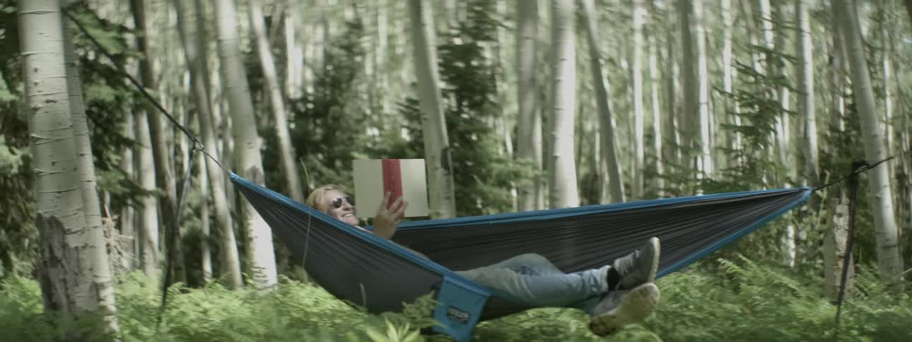 Man laying in hammock in forest is reading book and laughing