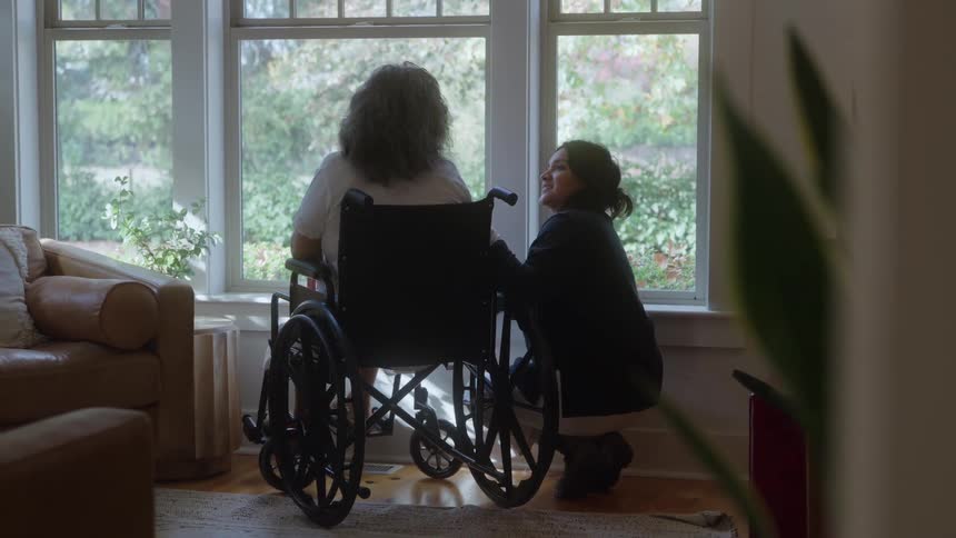 A dark haired woman kneels beside an older woman with gray curly hair sitting in a wheelchair both facing a large window overlooking a sunny outdoor scene in a cozy living room