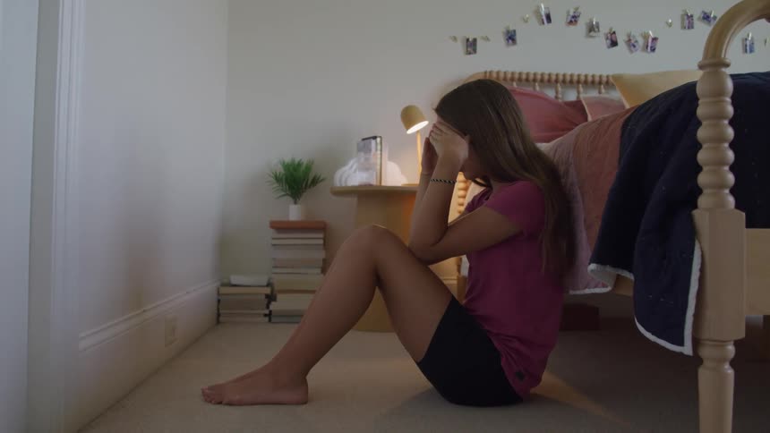 Distressed girl sitting on the bedroom floor with hands covering her face
