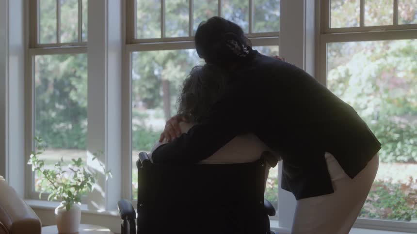 A woman walks up to an elderly person sitting in a wheelchair and hugs them