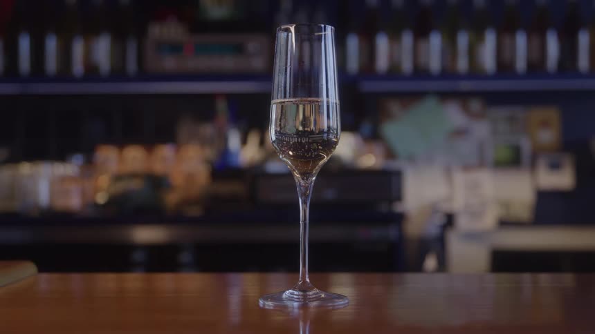 A tall flute of sparkling wine sits on a bar while a bartender moves in the blurred background