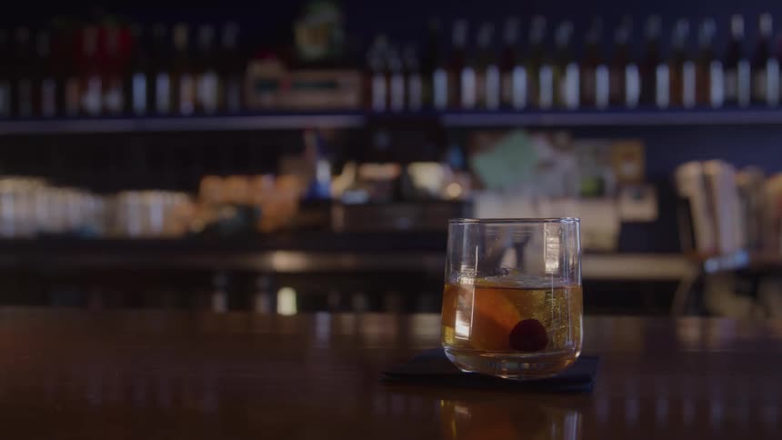 A hand places a short glass containing a whiskey liquid onto a wooden bar counter over a black napkin with rows of backlit liquor bottles visible in the background
