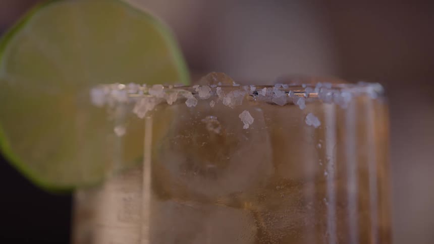 This is an extreme closeup of a beverage likely a margarita or a similar cocktail focusing on the rim of a clear textured glass the glass has a thick prominent salt rim and is garnished with a fresh lime wedge
