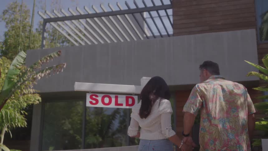 Homeowners stand next to a sold sign before taking each others hand and walking into their new home