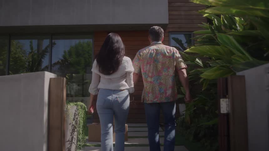 A man and a woman walk up a walkway toward the front of a modern house pass a large red sign and a stack of brown boxes and approach the front door surrounded by green tropical plants