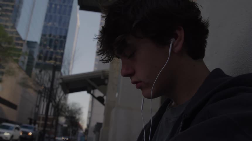 The video is a closeup shot of a young man likely a teen with dark curly hair sitting outdoors and looking downward with a pensive expressionhe is wearing white wired earbuds suggesting he is listening to music which adds to his sense of