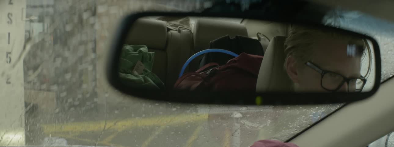 A driver's reflection is shown in a rearview mirror while it rains
