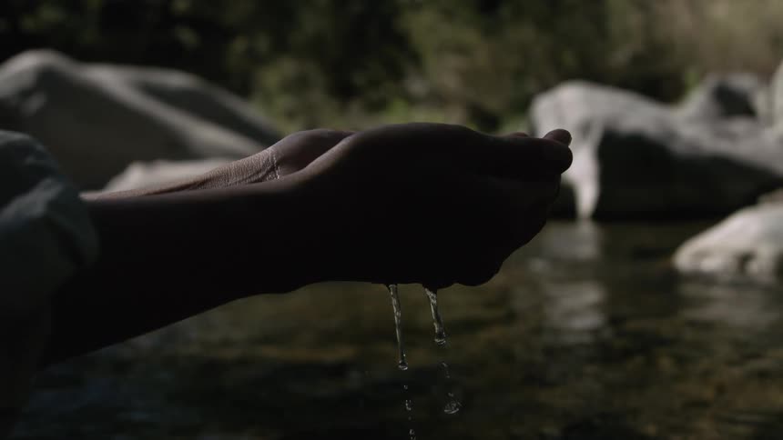 Water drips from the cupped hands of a man near a stream passing multiple boulders
