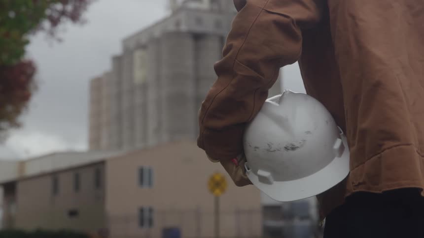 A person wearing a brown jacket walks in the foreground holding a white hard hat in the hand with a tall gray industrial building and a low structure visible in the background under a cloudy sky