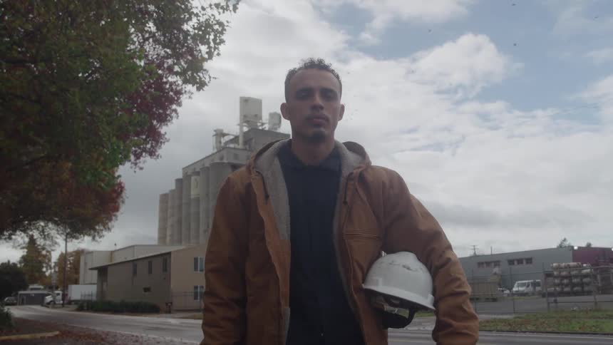 The video is an exterior shot of a man standing outdoors in a brown jacket and shirt holding a white hardhatbehind him is an industrial scene with a tall silo or grain elevator structure and other lowlying buildings beneath an overcast sky