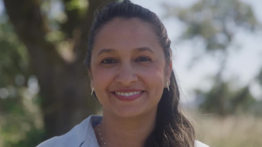 A woman with dark hair tied back turns her head from the side to the front and smiles at the camera wearing earrings and a collared shirt standing outdoors with blurred trees in the background