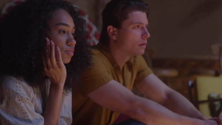 The video is a mediumclose shot of a young woman with long curly hair dressed in a lightcolored blouse seated and watching something offcamera with a thoughtful expressionshe is sitting beside a person whose arm is resting near her on a sofa
