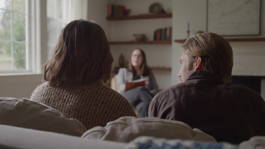 Husband and wife sitting on a couch listen to their counselor during a daytime session