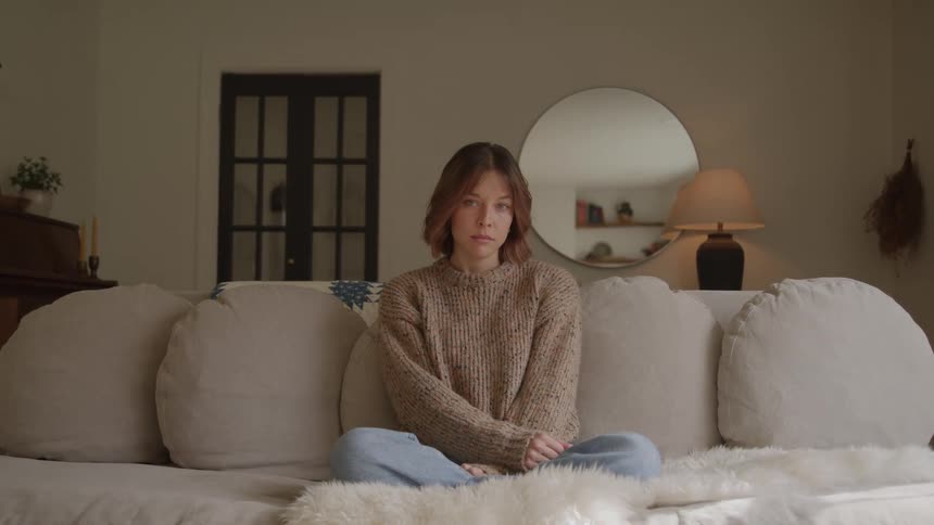 Young woman sits cross legged on a sofa in a dimly lit living room
