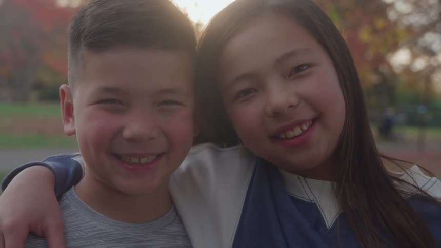 The video is a medium closeup of two children a boy and a girl standing close together outdoors in a parkthey are smiling brightly at the camera with their heads slightly tilted suggesting a moment of sibling affection or friendship against the bac