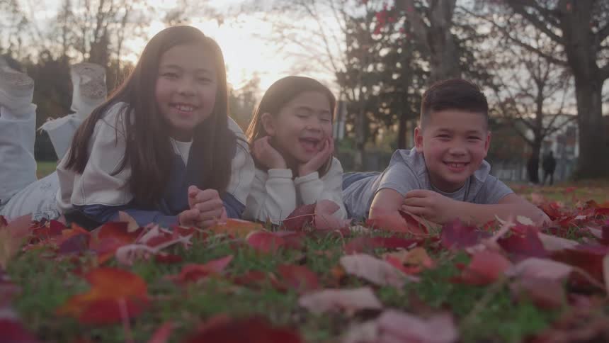 Three children lying on fall leaves in a park at sunset