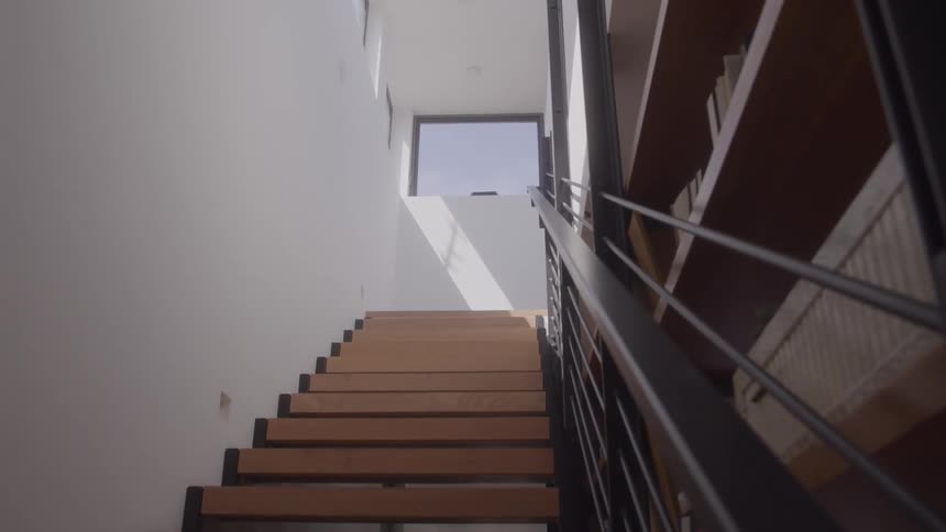 The video shows an upward view of a modern multistory staircase with wooden steps and a black metal railingthe staircase leads toward a small window at the top which is brightly lit by natural daylight